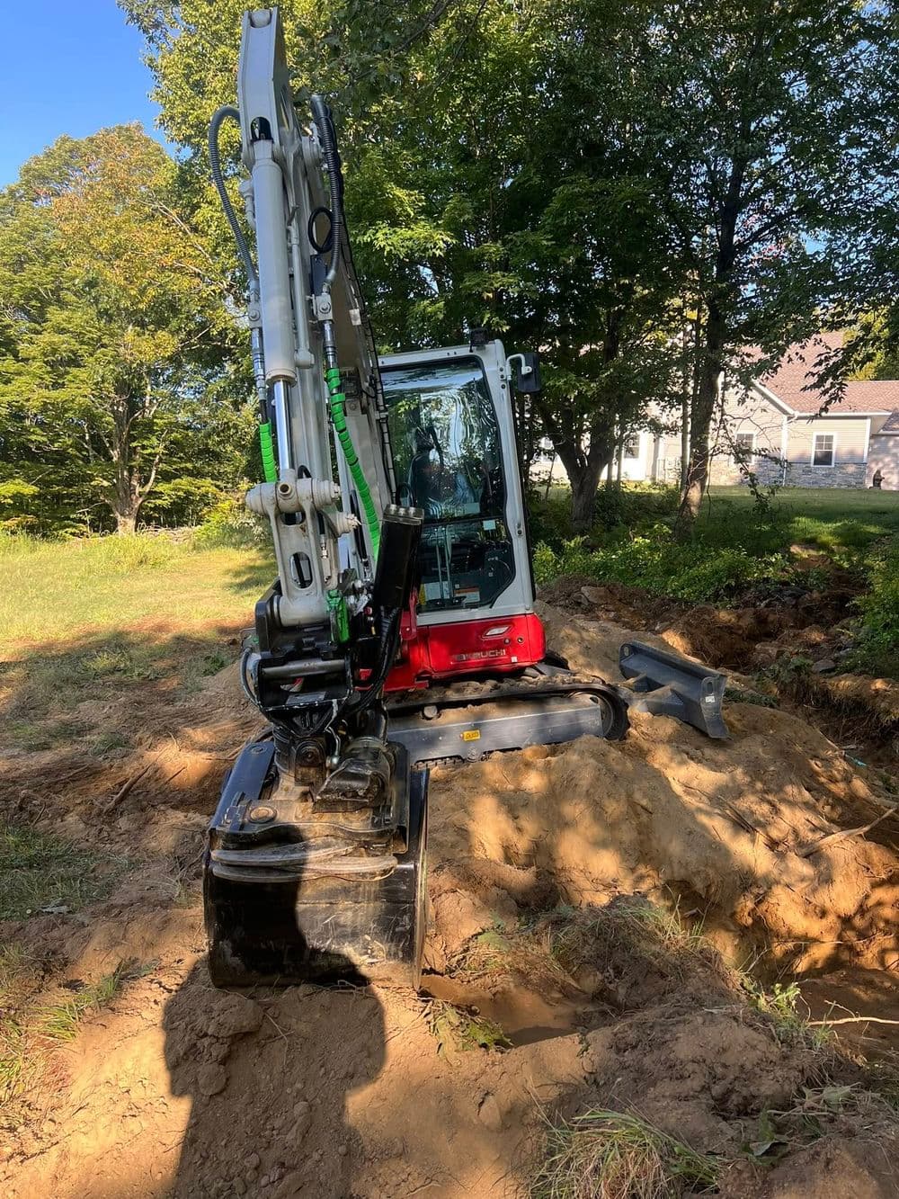 Excavator digging in a wooded area with a house in the background.