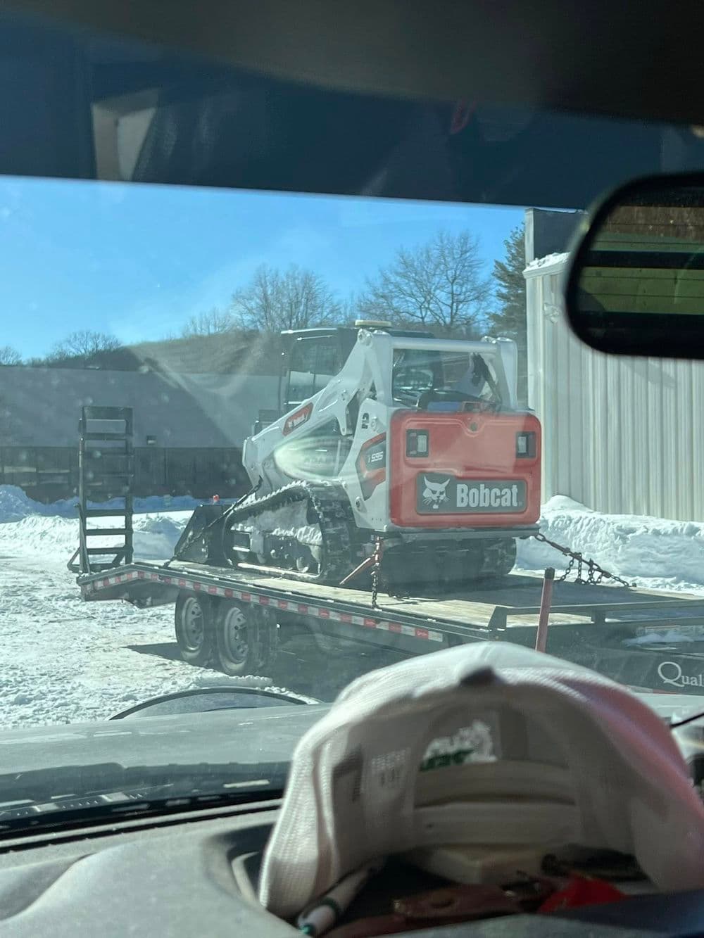Bobcat skid steer loader on a trailer in snowy landscape, viewed from a vehicle interior.