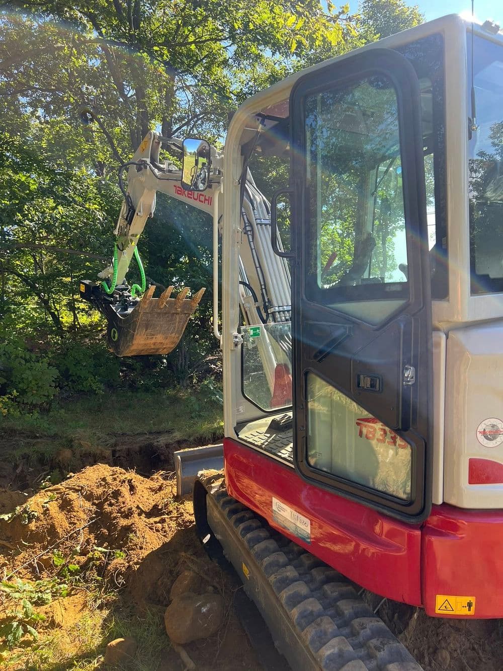 Mini excavator digging in soil, equipped with a claw attachment, surrounded by trees.